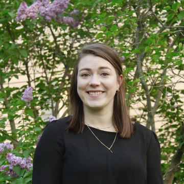 A smiling woman with dark hair in a black shirt standing in front of a lilac bush